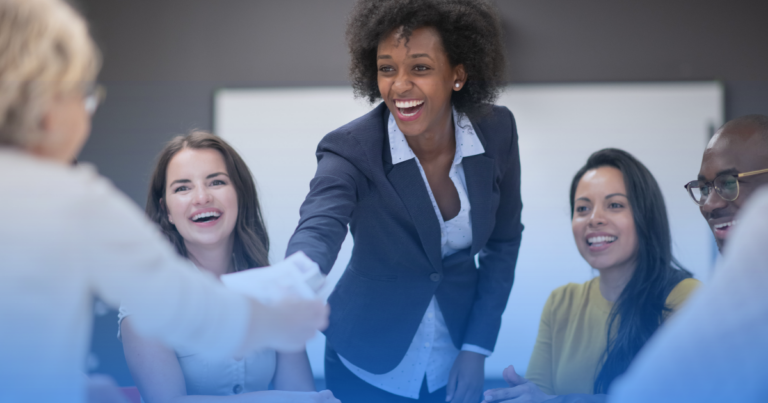Diverse Group of Coworkers Smiling As a Woman in a Navy Blazer Shakes Hands with a Colleague Across the Table